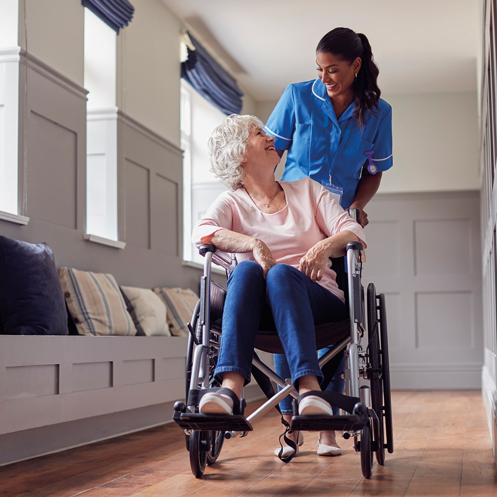 Female care worker in uniform pushes a senior woman in a wheelchair.
