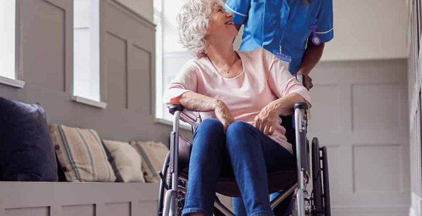 Female care worker in uniform pushes a senior woman in a wheelchair.